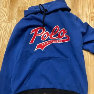 Blå hoodie från Ralph Lauren - Säljer en snygg blå hoodie från Ralph Lauren med stort rött Polo-tryck på framsidan. Den har en klassisk känguruficka och justerbar huva med svarta snören. Perfekt för en avslappnad stil eller kyliga dagar. Passar både till vardags och chillkvällar.