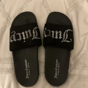 Svarta sandaler från Juicy Couture - Snygga svarta sandaler från Juicy Couture med glittriga detaljer på remmen. Perfekta för sommardagar eller en avslappnad dag på stan. De har en bekväm passform och är lätta att ta på och av. Ett måste för den som vill ha både stil och komfort!