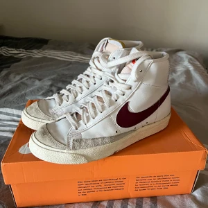 Nike Blazer Mid sneakers i vitt och vinrött - Säljer ett par Nike Blazer Mid sneakers i mycket bra skick. De är vita med en snygg vinröd swoosh och har klassisk snörning. Perfekta för både vardag och fest! Kommer i originalkartong.