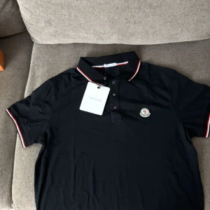 Svart pikétröja från Moncler - Säljer en snygg svart pikétröja från Moncler i nytt skick. Den har en klassisk krage med röd och vit kant samt Monclers logga på bröstet. Perfekt för både vardag och fest! 🖤