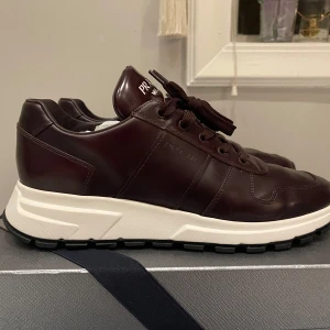 Prada skor, nypris 8k - Säljer ett par snygga bruna sneakers från Prada i skinn. De har en vit sula och klassisk snörning. Perfekta för både vardag och festliga tillfällen. Skorna är i mycket bra skick och kommer i originalförpackning.