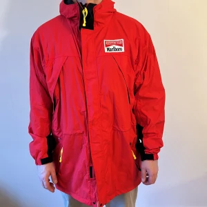 Retro Röd vindjacka från Marlboro - Säljer en cool röd vindjacka från Marlboro Adventure Team. Den har en dragkedja framtill och praktiska fickor med gula detaljer. Perfekt för höst och vår med sin långa passform och huva. Jackan är i bra skick och redo för nya äventyr!