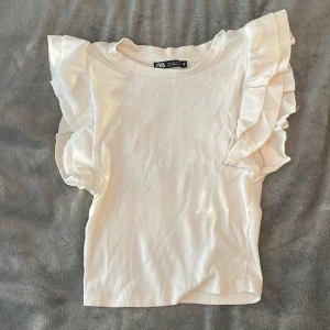 Vit ribbad topp från Zara - Säljer en superfin vit ribbad topp från Zara i storlek S. Den har volangdetaljer på ärmarna som ger en söt touch. Mycket bra skick