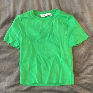 Grön ribbad topp från Zara - Säljer en snygg grön ribbad topp från Zara i storlek S. Den har en enkel design med korta ärmar och rund halsringning. Perfekt för både vardag och festliga tillfällen. Toppen är i mycket bra skick och redo för en ny garderob!
