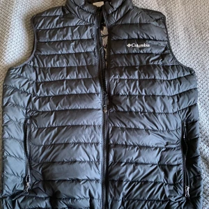 Columbia - Säljer en svart dunväst från Columbia i mycket bra skick. Västen har en dragkedja framtill och är quiltad för extra värme. Perfekt för höst och vår när du vill hålla dig varm utan att bära en jacka. Den har en stilren design och passar till de flesta outfits.