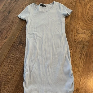 Ljusblå ribbad klänning - Säljer en superfin ljusblå ribbad klänning. Den är kortärmad och har en snygg rynkdetalj på sidan som ger en cool look. Perfekt för sommardagar eller en avslappnad vårdag. Klänningen är i bra skick och passar perfekt för en casual stil.