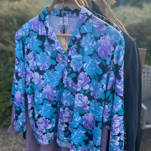 Blommig skjorta från Bloch Design - Snygg blommig skjorta från Bloch Design i härliga blå och lila nyanser. Perfekt för att liva upp garderoben med sitt färgglada mönster. Skjortan har en klassisk krage och långa ärmar, vilket gör den idealisk för både vår och sommar. Tillverkad i Danmark, vilket ger den en unik touch.