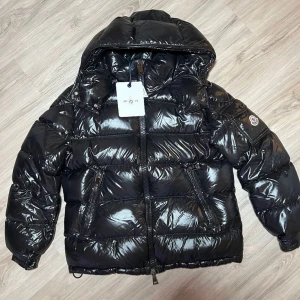 Svart glansig dunjacka från Moncler - Säljer en snygg svart glansig dunjacka från Moncler. Jackan har en pufferstil med dragkedja och huva, perfekt för kalla vinterdagar. Den är i mycket bra skick och har en stilren look som passar både till vardags och fest. Storleken är (S).