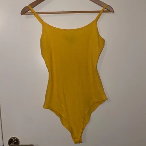 Gul ribbad body - Snygg gul body i ribbat material, perfekt för sommaren! Den har smala axelband och en enkel, stilren design som passar både för stranden och stan. Materialet är stretchigt och bekvämt, vilket ger en bra passform. Den är aldrig använd men lappen finns ej kvar. Storlek XS men stor i storleken så passar även någon med storlek M.