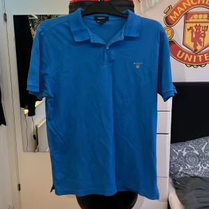 Blå pikétröja från GANT - Säljer en snygg blå pikétröja från GANT i storlek L. Tröjan har en klassisk krage och korta ärmar, perfekt för både vardag och lite finare tillfällen. Den är i bra skick och har GANT-loggan broderad på bröstet. Passar perfekt till sommaren eller våren! 💙