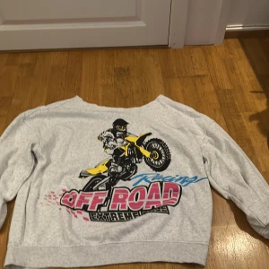 Grå sweatshirt med cross tryck - Säljer en grå sweatshirt med ett cross tryck på.  Med en skuren krage, off-shoulder. Materialet är en blandning av bomull och polyester, vilket gör den både bekväm och hållbar.