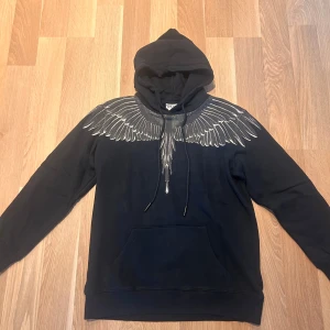 Marcelo Burlon Hoodie - Säljer en svart hoodie från Marcelo Burlon med ett coolt vingmönster i grått och vitt över axlarna. Perfekt för höst och vinter, den är både stilren och bekväm. Hoodien har en klassisk passform med känguruficka och justerbar huva. Passar perfekt för både vardag och fest!