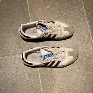 Säljer ett par klassiska Adidas Samba sneakers i mycket bra skick. De är vita med svarta ränder och har en snygg grå mockadetalj vid tån. Perfekta för både vardag och träning. Storlek är inte specificerad, men de ser ut att vara i herrstorlek. Skorna har snörning och en slitstark sula.