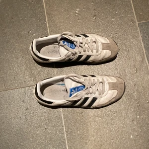 Adidas Samba sneakers - Säljer ett par klassiska Adidas Samba sneakers i mycket bra skick. De är vita med svarta ränder och har en snygg grå mockadetalj vid tån. Perfekta för både vardag och träning. Storlek är inte specificerad, men de ser ut att vara i herrstorlek. Skorna har snörning och en slitstark sula.