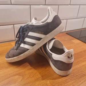 Adidas Gazelle - marinblå suede - Säljer ett par klassiska Adidas Gazelle i marinblå suede med vita detaljer och snörning. Skorna har en snygg retrostil med den ikoniska trefoil-loggan på hälen. Perfekta för både vardag, office eller fest!