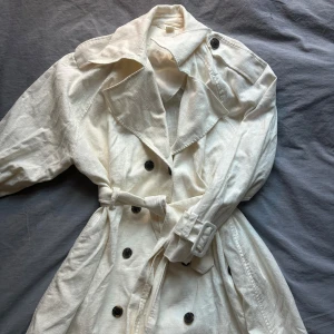 Vit Trenchcoat i Hampa Och Bomull - Säljer en stilren vit trenchcoat från H&M gjord i bomull och hampa blandning vilket är en mer klimatvänlig textil och ger en värmehållande men luftig känsla lik till linen (obs: inte faktisk linen) Köpt för ca 4 år sedan för 800kr, använd endast 2-3 gånger utan defekter Säljer då den ej kommer till användning