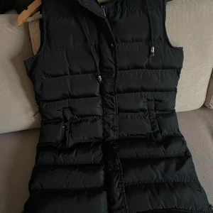 Svart dunväst med huva - Säljer en snygg svart dunväst i mycket bra skick. Västen har en praktisk huva och dragkedja framtill. Perfekt för höst och vinter när du vill hålla dig varm och bekväm. Den har en stilren design som passar till det mesta. Hör av dig vid intresse! (Kan mötas upp i Stockholm) 💕