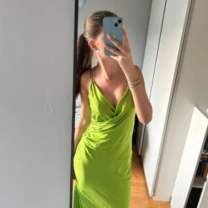 Grön omlottklänning - Säljer en snygg grön omlottklänning i ett mjukt och glansigt material. Klänningen har smala axelband och en elegant drapering framtill. Perfekt för sommarens fester eller en kväll ute. Den är i mycket bra skick och ger en fräsch och stilren look.