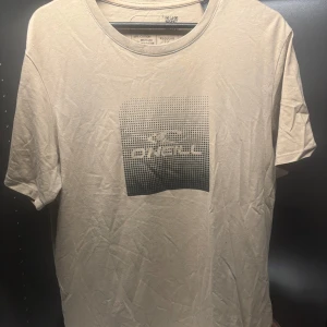 Beige t-shirt från O'Neill - Säljer en beige t-shirt från O'Neill med ett coolt svart tryck på framsidan. Tillverkad av 60% bomull och 40% återvunnen polyester. Perfekt för en avslappnad stil.