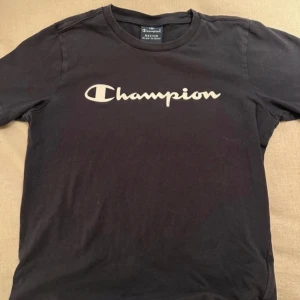 Svart t-shirt från Champion - Snygg svart t-shirt från Champion med deras ikoniska logga tryckt på bröstet. Perfekt för en avslappnad stil. Tillverkad i mjukt material för extra komfort.
