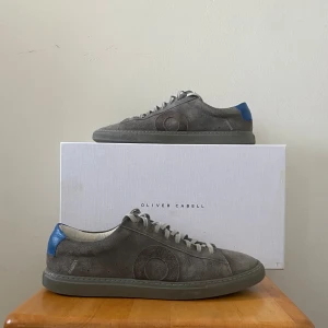 Oliver Cabell skor - Snygga grå sneakers från Oliver Cabell med blå detalj på hälen. Skorna har en stilren design med snörning och är tillverkade i mocka. Perfekta för våren och sommaren. Storleken är 40-41. De är precis som nya och knappt använda.