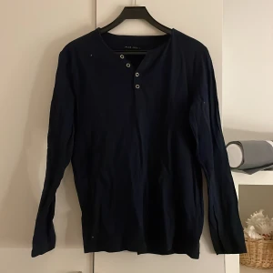 Mörkblå långärmad tröja från Pier One - Säljer en stilren mörkblå långärmad tröja från Pier One. Tröjan har en knappdetalj vid halsen som ger en avslappnad look. Perfekt för en casual stil.