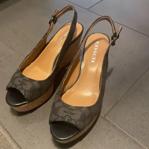 Svarta kilklackar från Coach - Snygga svarta kilklackar från Coach med öppen tå och justerbar rem runt hälen. Skorna har ett diskret mönster och en bekväm korksula som ger extra höjd. Perfekta för en stilren look.