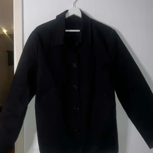 Svart rock från Stutterheim - Snygg svart rock från Stutterheim med knappar framtill. Perfekt för en stilren look. Rocken har långa ärmar och en klassisk krage. Passar bra till både vardag och mer formella tillfällen.