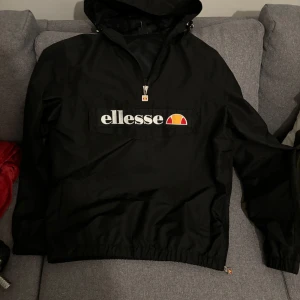 Svart vindjacka från Ellesse - Säljer en svart vindjacka från Ellesse med stor logga på framsidan. Jackan har en dragkedja vid halsen och en praktisk huva. Perfekt för blåsiga dagar och har en sportig look. Passar bra till både träning och vardag.