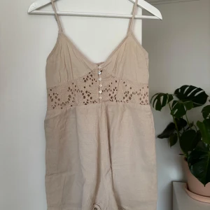 Beige linnebyxdress med spetsdetaljer - Säljer en söt beige byxdress med smala axelband och v-ringning. Byxdressen har fina spetsdetaljer och knappar framtill. Perfekt för varma sommardagar! 🌞