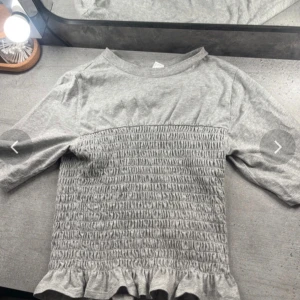 Grå tröja från Divided - Säljer en grå tröja från Divided med smockdetaljer och volangkant. Tröjan har trekvartsärmar och en rund halsringning. Perfekt för en avslappnad stil. 🩶 Superfin , bara testad knappast andvänd 