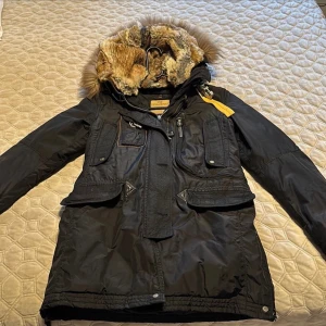 Svart vinterjacka från Parajumpers - Säljer en svart vinterjacka från Parajumpers med pälsfodrad huva och flera praktiska fickor. Jackan har en robust dragkedja och knappar framtill samt en gul detalj vid axeln. Perfekt för kalla dagar! 🖤