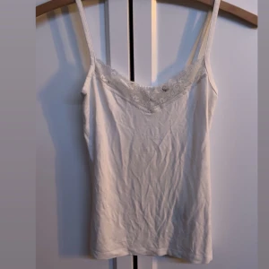 Vit linne med spetsdetaljer - Säljer ett stilrent vitt linne med smala axelband och vacker spetsdetalj längs halsringningen. Perfekt för lager-på-lager eller som det är under varma dagar. Säljer för jag har flera likadana, simpelt jätte snyggt linne. 🤍