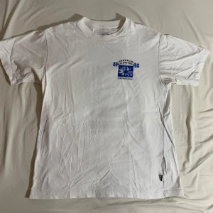 Vit T-Shirt från Adidas Skateboarding - Säljer en vit t-shirt från Adidas med ett coolt blått tryck på både fram- och baksidan. Framsidan har en liten logga medan baksidan har ett större tryck med texten 'Brooklyn Flowers'. Perfekt för en avslappnad stil. Köpt från Urban Outfitters. 