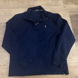 Ralph Lauren Quarter Zip - Säljer en stilren mörkblå tröja från Polo Ralph Lauren med dragkedja vid halsen och det klassiska logotypbroderiet på bröstet. Perfekt för en avslappnad men ändå elegant look. Tröjan har långa ärmar och är tillverkad i ett mjukt material.