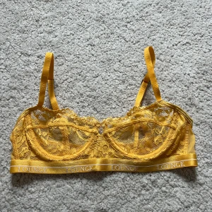 Gul spets-BH från Lounge - Säljer en snygg gul spets-BH från Lounge med justerbara axelband och hakknäppning bak. BH:n har en elegant spetsdesign och ett elastiskt band med Lounge-loggan. Perfekt för att känna sig både bekväm och stilren.