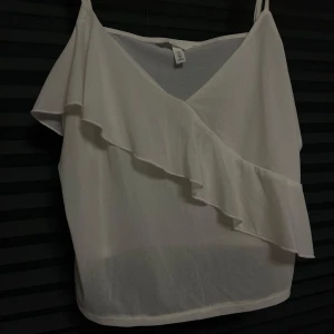Vit linne med volangdetalj - Snygg vit linne från H&M med volangdetalj över bröstet. Perfekt för varma sommardagar med sin lätta och luftiga design. Axelbandslös stil som ger en elegant touch.