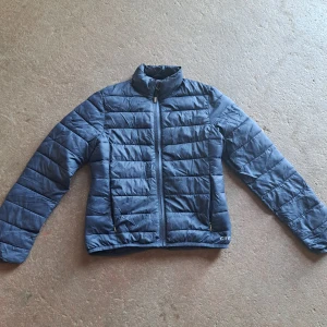 Blå pufferjacka från CRW - Säljer en blå pufferjacka från CRW, perfekt för kyliga dagar. Jackan har en dragkedja framtill och långa ärmar. Den quiltade designen ger extra värme och stil. Passar bra för både vardag och utomhusaktiviteter.