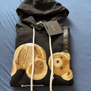 Svart hoodie med nallemotiv från Palm Angels - Säljer en svart hoodie från Palm Angels med ett unikt nallemotiv på framsidan. Hoodien har en justerbar dragsko i huvan och är tillverkad i mjukt material för extra komfort. Perfekt för en avslappnad stil. 🖤🐻Tröjan är helt ny och oanvänt förutom att jag har testat den.