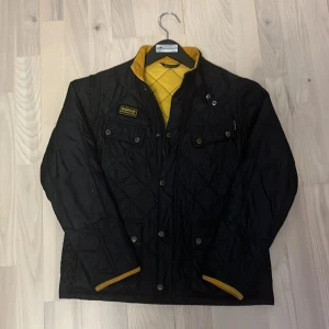 Svart quiltad jacka från Barbour - Säljer en snygg svart quiltad jacka från Barbour med gul insida. Jackan har långa ärmar, knappar och en dragkedja framtill. Den har också två bröstfickor med knappar och en broderad Barbour-logga på bröstet. Perfekt för höst och vår! Steal!