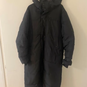 Svart lång dunjacka med huva - Säljer en svart lång dunjacka med huva. Jackan har en dragkedja som är dock sönder men det finns knappar framtill och långa ärmar. Perfekt för kalla dagar när du vill hålla dig varm och bekväm.
