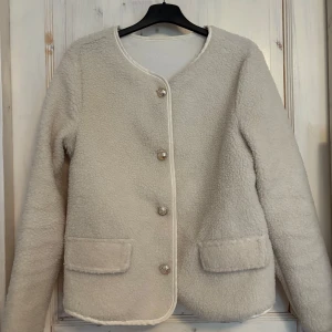Vit teddyjacka med knappar - Mysig vit teddyjacka med rund hals och dekorativa knappar framtill. Jackan har två praktiska fickor och en stilren design. Perfekt till våren endast använt en gång