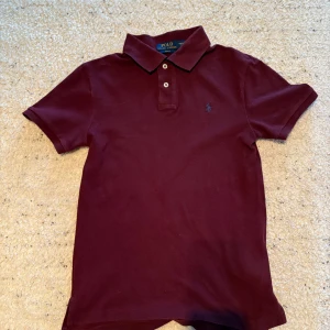 Vinröd pikétröja från Ralph Lauren - Säljer en stilren vinröd pikétröja från Ralph Lauren. Tröjan har en klassisk krage med två knappar och en broderad logotyp på bröstet. Perfekt för en avslappnad men ändå elegant look. Passar bra till både jeans och chinos.