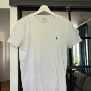 Vit t-shirt från Ralph Lauren - Säljer en klassisk vit t-shirt från Ralph Lauren med korta ärmar och en broderad logga i blått på bröstet. Perfekt för en stilren och avslappnad look. Passar bra till jeans eller shorts.
