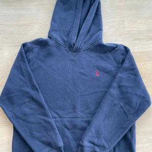 Mörkblå hoodie från Ralph Lauren - Säljer en snygg mörkblå hoodie från Ralph Lauren med det klassiska röda broderade logotypen på bröstet. Tröjan har en bekväm passform med långa ärmar och en praktisk huva. Perfekt för en avslappnad stil. Storlek 170cm/15years