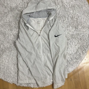 Vit vindjacka från Nike - Säljer en vit vindjacka från Nike med ett diskret rutmönster och svart logga på bröstet. Jackan har dragkedja och huva, perfekt för sportiga aktiviteter. Den är tillverkad i syntetmaterial och har en stilren design.