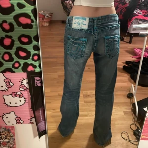 Blå jeans med broderade bakfickor - Snygga blå jeans från True Religion med unika turkosa broderier på bakfickorna. De har en bootcut-stil och låg midja, vilket ger en avslappnad och trendig look. Perfekta för dig som vill sticka ut med detaljerade jeans.💓