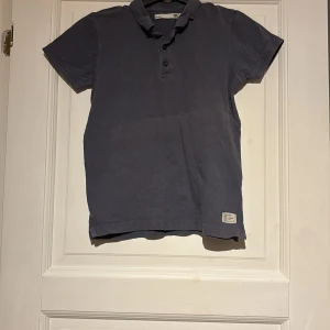 Mörkblå pikétröja - Säljer en stilren mörkblå pikétröja med krage och knappar framtill. Perfekt för en avslappnad men ändå snygg look. Tröjan har korta ärmar och en liten etikettdetalj vid nederkanten. Det är två likadana för de priset i samma paket.