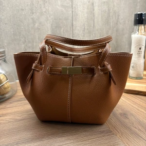 Brun axelväska med dekorativt spänne - Snygg poppis brun axelväska i syntetmaterial med ett dekorativt spänne framtill. Väskan har en praktisk axelrem och stilrena handtag. Perfekt för att bära dina dagliga nödvändigheter med stil.