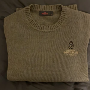 stickad tröja från Morris - Säljer en stilren olivgrön stickad tröja från Morris. Tröjan har en broderad logotyp på bröstet och ribbade kanter vid hals, ärmslut och nederkant. Perfekt för en avslappnad men ändå elegant look. Tröjan är i nyskick använd max 3 gånger. Passar dig mellan 180-185 ish beroende på vikt också.